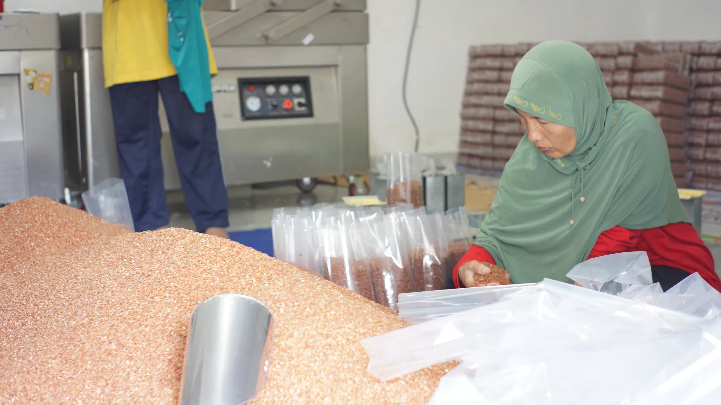 Pekerja Sirtanio mengisi kemasan plastik dengan beras merah di samping mesin vakum, dengan tumpukan beras di latar depan.